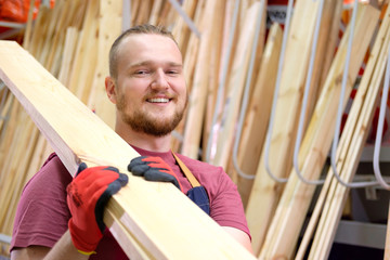 Smiling salesman or seller in construction store or warehouse wood section holding a pack of...