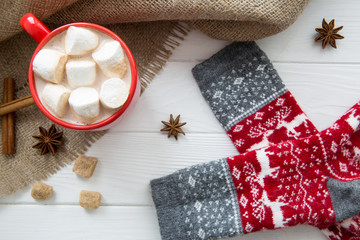 Christmas red cup with hot chocolate and marshmallow. Socks with christmas ornament and deer. Hot cocoa with milk, anise and cinnamon on white wooden rustic background, hessian (sacking) top view.