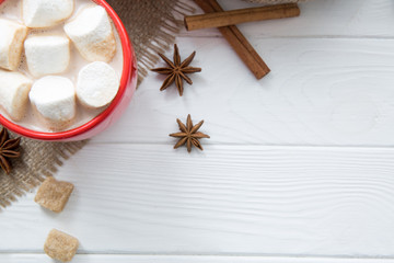 Christmas red cup with hot chocolate and marshmallow. Hot cocoa with milk, anise and cinnamon rolls on white wooden rustic background, hessian (sacking) top view. Copy space.