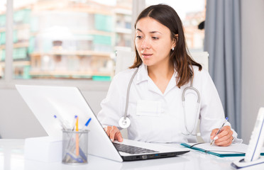 Attentively female doctor  working at laptop and writing papers