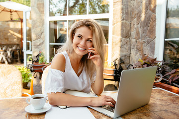 Laughing blonde businesswoman having a funny phone conversation, during working on laptop computer,...