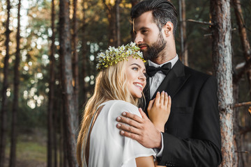 beautiful smiling bride in white dress and handsome groom in suit hugging in forest