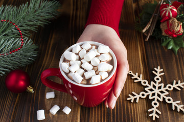 Obraz na płótnie Canvas Woman hand is holding the cup of hot chocolate with marshmallows, snowflake on the brown wooden background.
