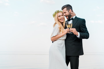 happy groom and bride hugging and clinking with glasses of champagne on beach