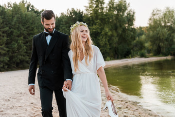wedding couple holding hands and walking on beach, laughing bride holding high heels in hand