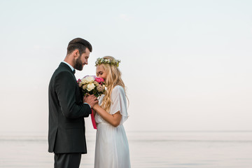 side view of wedding couple standing with bouquet on beach, bride sniffing roses