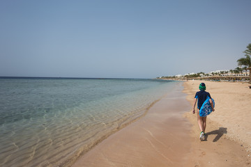 Cute white tanned kid dressed in swimsuit, cap and aqua shoes having fun at summer sandy beach using blue inflatable surf board. Boy walking alone with his back to camera. Horizontal color photography