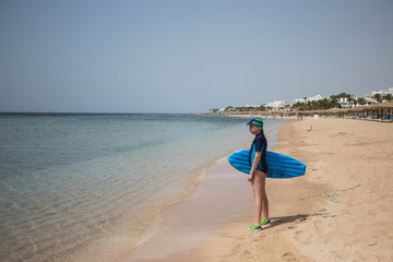 Cute white tanned kid dressed in swimsuit, cap and aqua shoes having fun at summer sandy beach using blue inflatable surf board. Horizontal color photography.