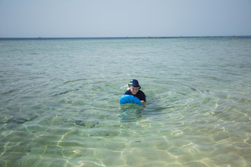 Cute white tanned kid in swimsuit, cap and aqua shoes having fun at summer sandy beach using blue inflatable surf board. Horizontal color photography.