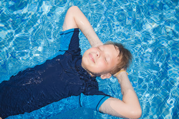 Portrait of cute calm blond white kid relaxing in clear blue water of outdoors sunny swimming pool lying calmly on back. Horizontal color photography.