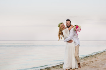 happy bride and groom hugging on beach and holding wedding bouquet