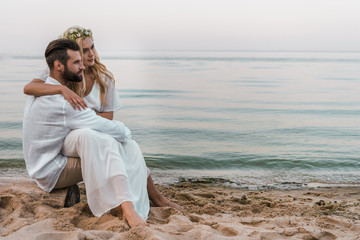 elegant groom and bride sitting on log on beach and looking away