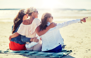 summer vacation, holidays, travel and people concept - group of smiling young women in sunglasses sitting on beach blanket and pointing finger to something