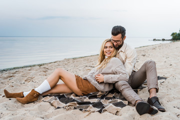 smiling couple in autumn outfit sitting on blanket and hugging on beach
