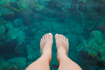 Closeup top view of female pedicured legs under crystal blue water. Woman sits on pontoon at hotel resort enjoying watching many colorful fish swimming around feet. Point of view horizontal photo.