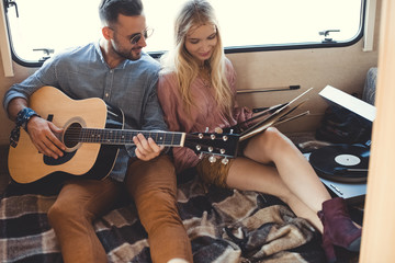 happy musician playing on acoustic guitar while girl holding vinyl record inside campervan