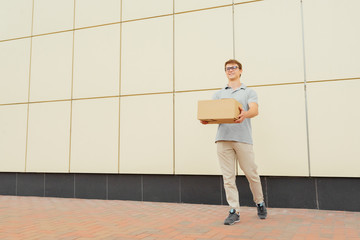 Cheerful attractive courier in eyeglasses, looking at camera while holding a delivery box and clipboard, standing against the wall, outdoors.