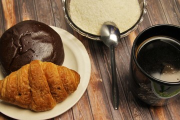 A cup of black coffee, a plate of pastry, and a bowl of sugar on a brown wooden background