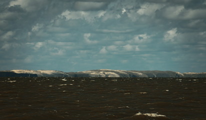 Sea water with waves and land with sand dunes in horizon with clouds