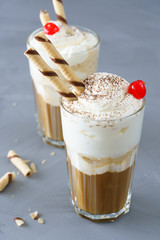 Ice coffee topped with vanilla ice cream, whipped cream, wafer straws and cocktail cherry. Gray table, high resolution.