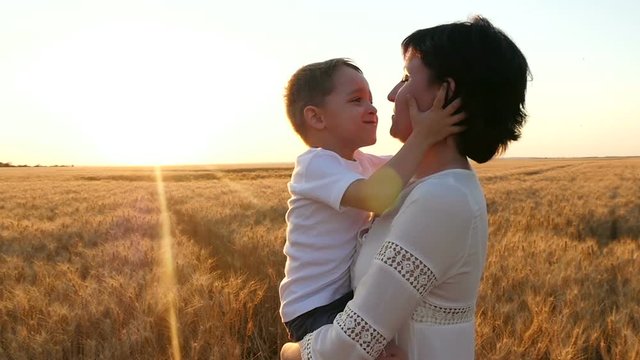A happy mother holds a child in her arms in a field of wheat. Happy child kisses mother's face, mother kisses child on sunset background.