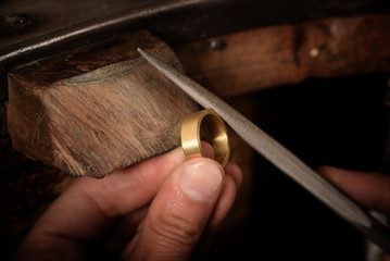 goldsmith hand holds a golden ring on the wooden workbench and works on it with a metal file, close up with copy space