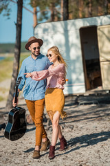 young hippie couple hugging and walking with guitar near campervan, girl pointing somewhere