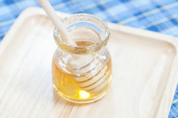 Open glass jar of liquid honey from honey dipper on wood pan and fabric pattern background