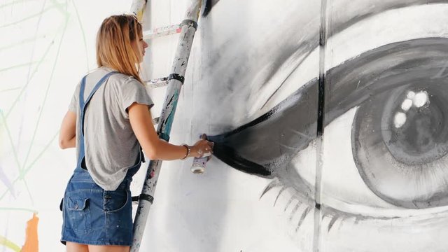 Beautiful girl making graffiti of big female face with aerosol spray on urban street wall. She standing on ladder. Creative art. Talented student in denim overalls drawing picture