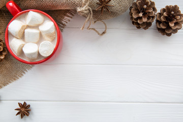 Christmas red cup with hot chocolate and marshmallow. Hot cocoa with milk, anise, cone decoration on white wooden rustic background, hessian top view. Copy space.
