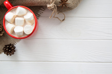 Christmas red cup with hot chocolate and marshmallow. Hot cocoa with milk, anise, cone decoration on white wooden rustic background, hessian top view. Copy space.