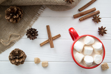 Christmas red cup with hot chocolate and marshmallow. Hot cocoa with milk, anise and cinnamon rolls, pine cone on white wooden rustic background, sacking top view.