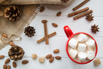 Christmas red cup with hot chocolate and marshmallow. Hot cocoa with milk, anise and cinnamon sticks, almonds and pine cones on white wooden rustic background, hessian, top view.