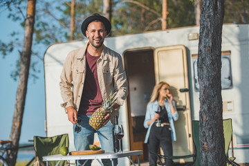 selective focus of happy man cutting pineapple while woman standing at campervan behind