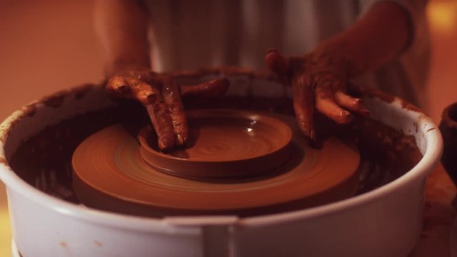 Rotating potter's wheel with woman's hands playfully working on it using fingers for shaping small brown clay dish with wide edges in dark light studio, having tray table with pottery tools.