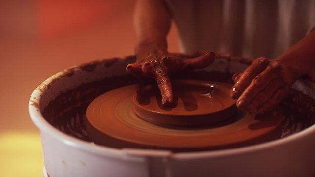 Side view of woman's hands professionally working on rotating pottery wheel forming bottom surface of small brown clay plate with wide edges in dark light studio.