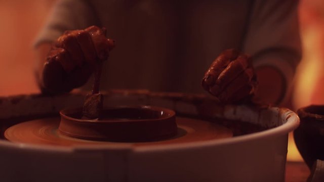 Side view of rotating potter's wheel with woman's hands professionally working on it using pottery tool to shape walls of small brown clay pot with wide edges in dark light studio.
