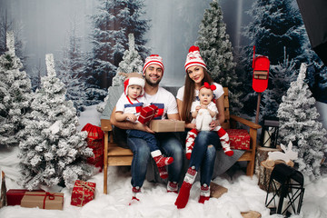Studio portrait of cheerful caucasian family in Santa hats with kids sitting on their knees holding Christmas presents with snowed fir trees around them. Looking at camera and smiling happily.