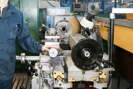 A Man Working In A Robe, Overalls Stands Next To An Industrial Lathe For Cutting, Turning Knives From Metals, Wood And Other Materials, Turning, Making Details And Spare Parts At The Factory