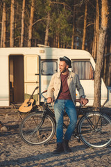 Fototapeta na wymiar young handsome man leaning on bicycle near campervan in forest