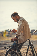 Obraz premium young handsome man using smartphone and leaning on bicycle outdoors