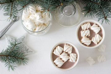 Cocoa with marshmelow in white cups and marshmallows in a glass jar on a white table with spruce branches and garland.