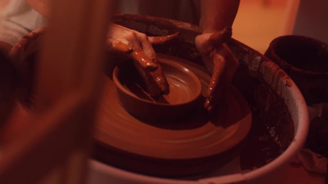 Camera showing woman in white sweater who is professionally working on potter's wheel with hands forming and shaping brown thin clay pot with wide edges in dark light studio.