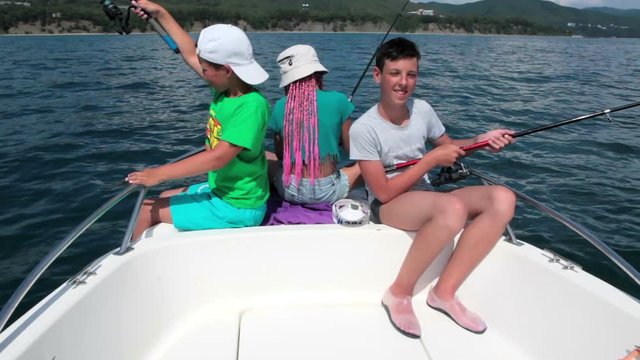 Three happy teen age kids sitting in boat with spinning tackles, have fishing excursion