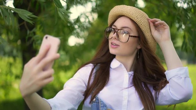 Hipster young woman with long red hair wearing glasses, a white shirt and a straw hat is making a selfie in a park on a summer day. Handheld medium shot