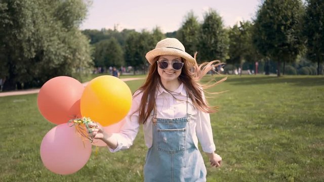 Young woman with long red hair and straw hat holding colored balloons, running towards the camera in a summer park. She is taking the hat off. Happiness and joy concept. Tracking medium shot