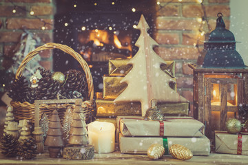 Christmas setting, wooden ornaments, presents and pine cones, tree on the table in front of fireplace with woodburner, star lights and garlands, lit lantern, created snow, selective focus, toned