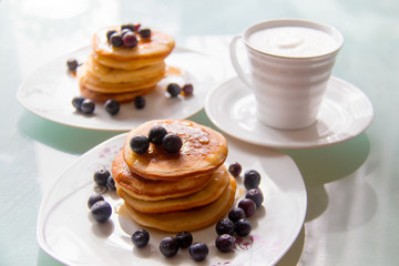 Breakfast pancakes on the stylish plate served with fresh blueberries and topped with original maple syrup, next cup of creamy white coffee on the glass table