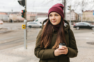 A girl drinks coffee from a disposable cup on the street in Prague in cool weather. The usual urban everyday life.
