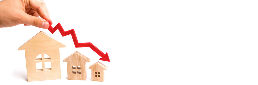 The hand holds a red arrow above the wooden houses down. The houses are decreasing. The concept of falling demand and supply in the real estate market
